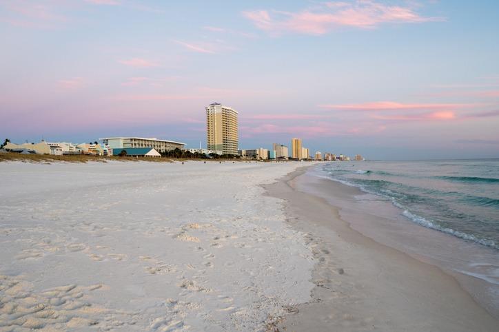 Panama City Beach rentals beachfront view with soft white sand, calm emerald waters, and high-rise resorts along the shoreline at sunset.